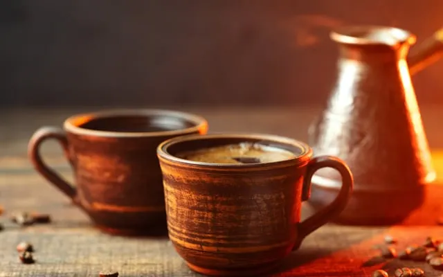 Two clay cups of coffee beside a copper coffee pot, with coffee beans on a wooden table
