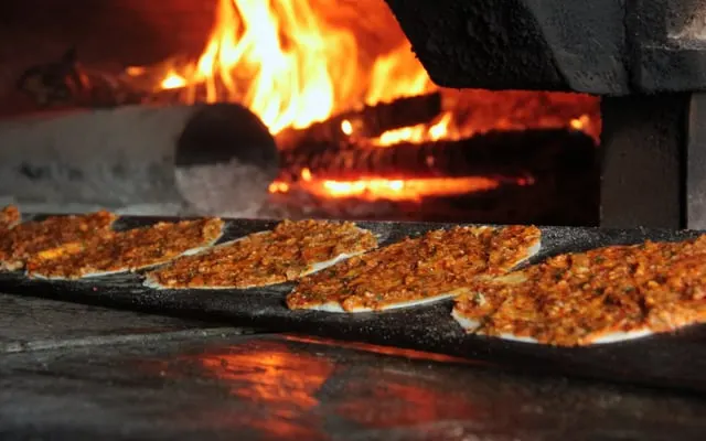 Thin flatbreads with a minced topping lined up on a baking peel in front of a wood fire