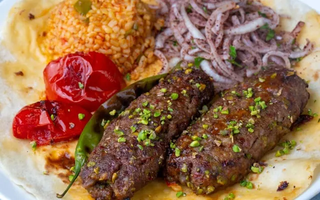 Two grilled minced-meat skewers on flatbread with bulgur, sumac onions and roasted tomatoes, topped with pistachio