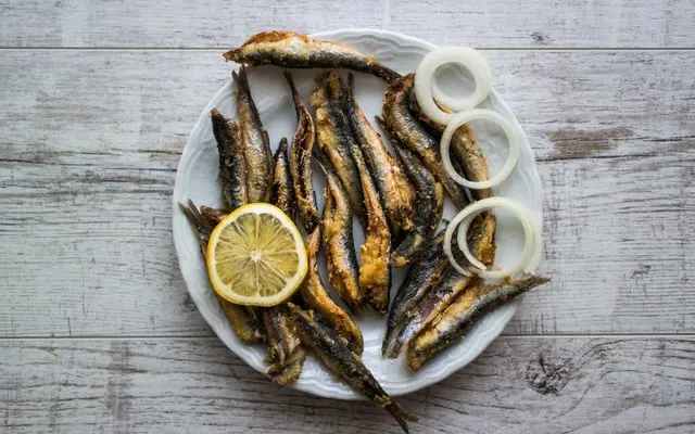 Plate of fried small fish with a lemon half and onion rings on a pale wooden table
