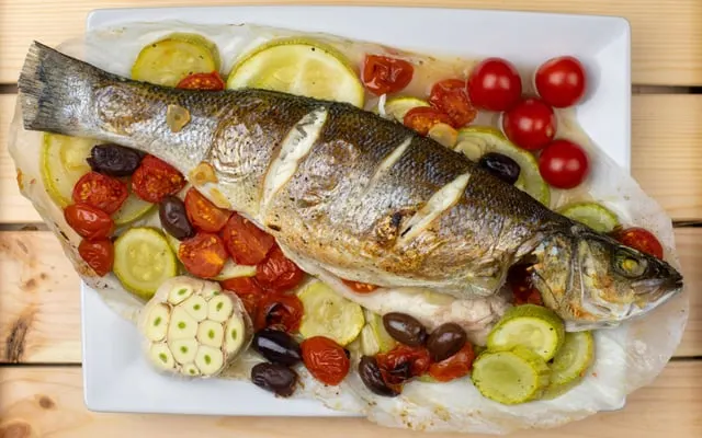 Whole roasted sea fish on baking paper with cherry tomatoes, courgette slices, olives and garlic