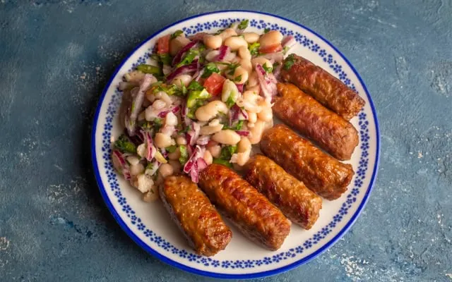 Grilled elongated meatballs on a patterned plate with a white bean, onion and parsley salad