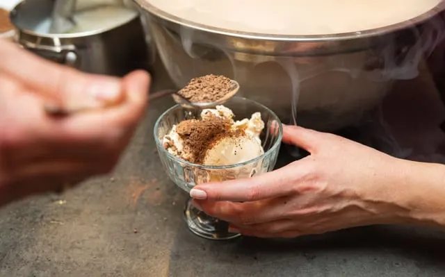 Ice cream in a glass dish being dusted with cocoa from a spoon, with vapour rising from a bowl behind