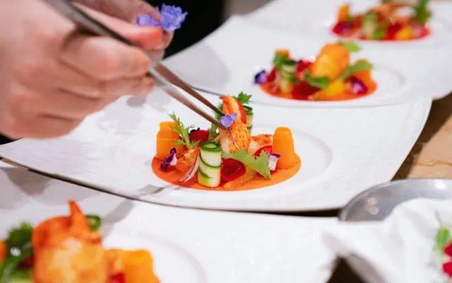 Chef placing a small flower with tweezers on a plated shellfish dish with carrot and cucumber