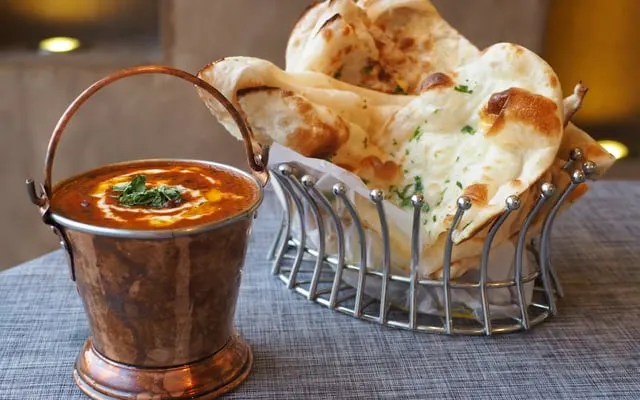 Copper bowl of creamy tomato curry next to a wire basket of naan bread