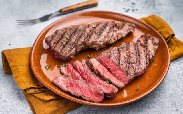 Grilled steak, partly sliced, on a terracotta plate with herbs and a mustard-coloured napkin