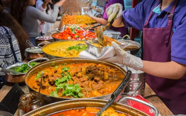 Street food stall worker in gloves serving curry from large pans to a queue of customers