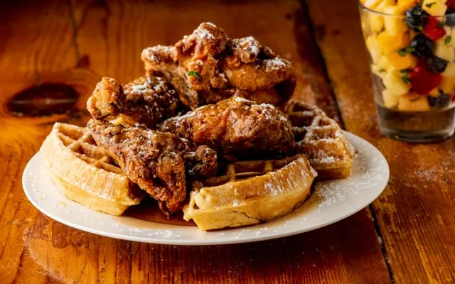 Fried chicken pieces on waffles dusted with sugar, with a glass of fruit salad beside the plate