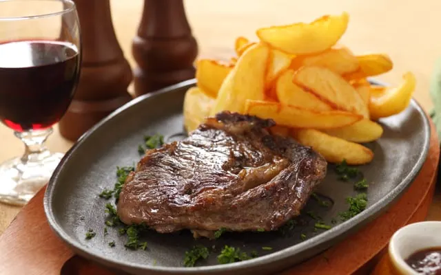 Grilled steak on a cast-iron plate with thick potato wedges and a small dish of sauce