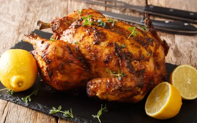 Whole roast chicken with herbs on a slate board, served with lemon halves and carving utensils