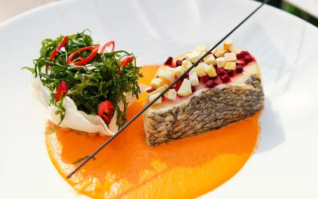 Plated fish fillet topped with diced garnish on orange sauce, beside a crisp chilli-topped herb salad