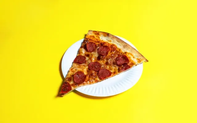 Slice of pizza topped with round red meat slices on a white paper plate against a yellow background